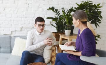 candid shot young man glasses talking about his problems during psychological therapy session sitting coach while mature female psychologist with copybook listening him making notes