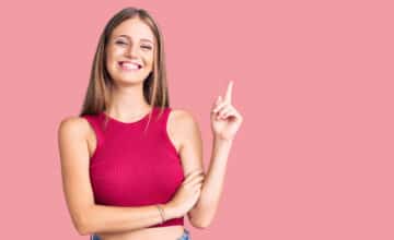 young beautiful blonde woman wearing elegant summer shirt with big smile face pointing with hand finger side looking camera